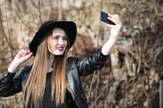 Giovane bella ragazza con un cappello e con un trucco scuro all'esterno. Ragazza in stile gotico per strada. Una ragazza cammina per la strada della città in un gilet di pelle con il telefono.