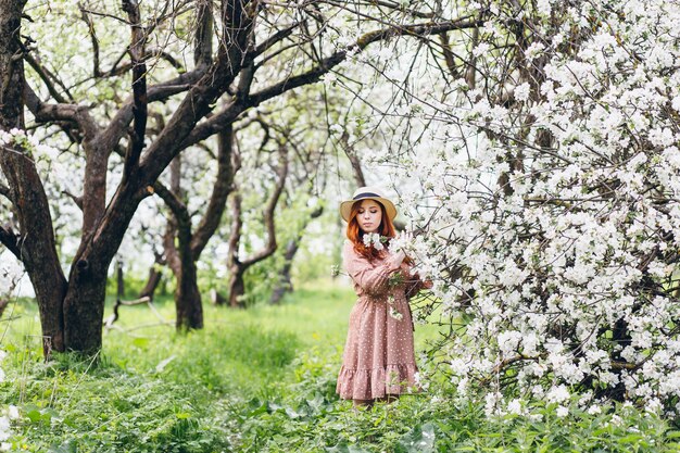 Giovane bella ragazza dai capelli rossi cammina in un frutteto di mele in fiore primaverile