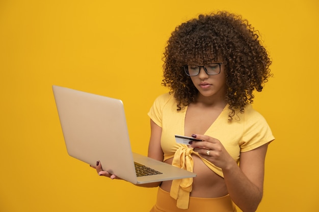 Giovane bella ragazza dei capelli ricci con il computer portatile e la carta di credito su fondo giallo, donna di affari che compera online.