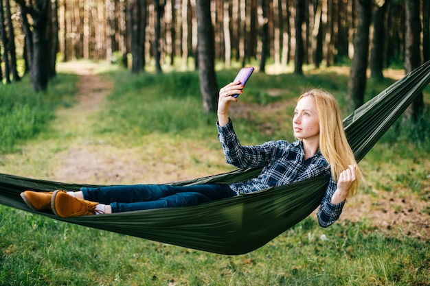 Giovane bella ragazza di rilassamento bionda in amaca e fare selfie nella foresta di estate