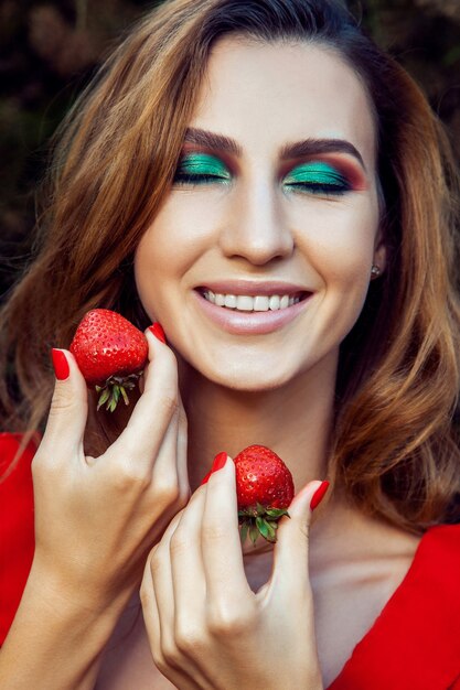 Giovane bella ragazza divertente felice con il vestito rosso e trucco che tiene fragola in estate nel parco. stile di vita sano, dieta bellezza e concetto di felicità.