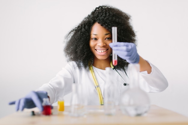 Giovane bella ragazza dottore in camice bianco con uno stetoscopio. seduto a un tavolo con boccette di reagenti su sfondo bianco. Assistente di laboratorio femminile con analisi del sangue per l'AIDS