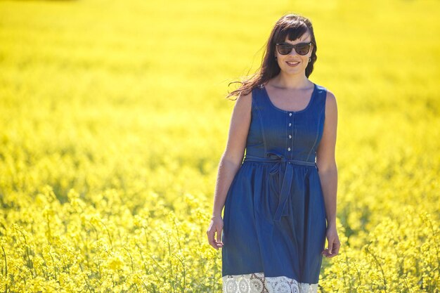 Giovane bella ragazza felice in un vestito blu con occhiali da sole sul campo di colza in fiore in estate