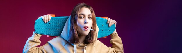 Giovane bella ragazza hipster alla moda vestita con una felpa con cappuccio tiene uno skateboard nelle sue mani su uno sfondo di studio viola Mangia un lecca-lecca