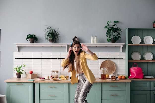 Giovane bella ragazza in abiti alla moda che si diverte in cucina Modello bruna felice con i capelli lunghi che ascolta musica e balla Donna alla moda in giacca gialla e cuffie blu che cantano