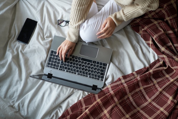 Giovane bella ragazza in maglione accogliente a letto a casa, lavorando su un computer portatile