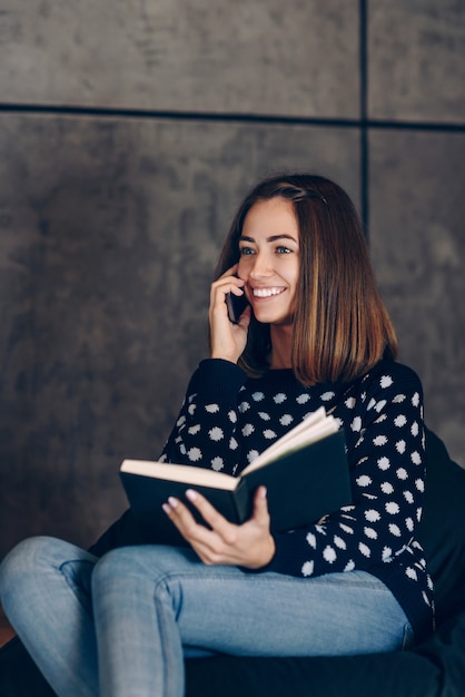 Giovane bella ragazza in maglione caldo, seduto con sketchbook e parlando al cellulare di un muro di cemento