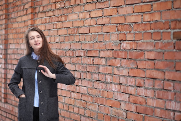 Giovane bella ragazza in un cappotto vicino alla posa del muro di mattoni rossi