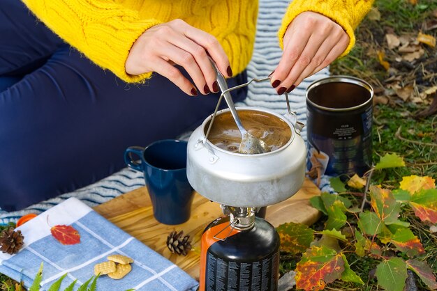 Giovane bella ragazza in un maglione giallo fa il caffè nella foresta su un bruciatore a gas. Preparare il caffè su un fornello primus nella foresta d'autunno, passo dopo passo