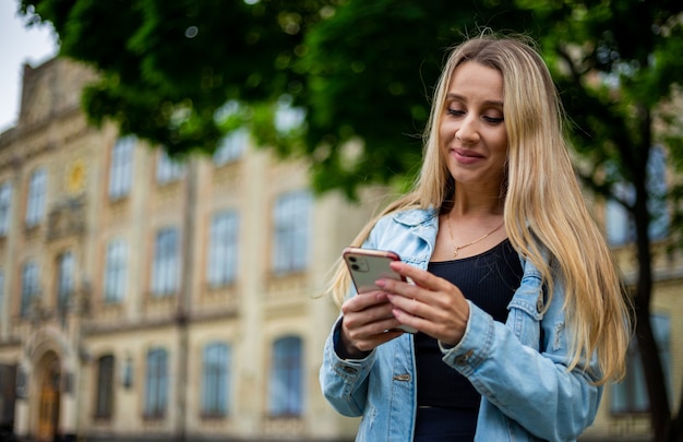 Giovane bella ragazza moderna alla moda in una giacca di jeans per strada guardando il telefono