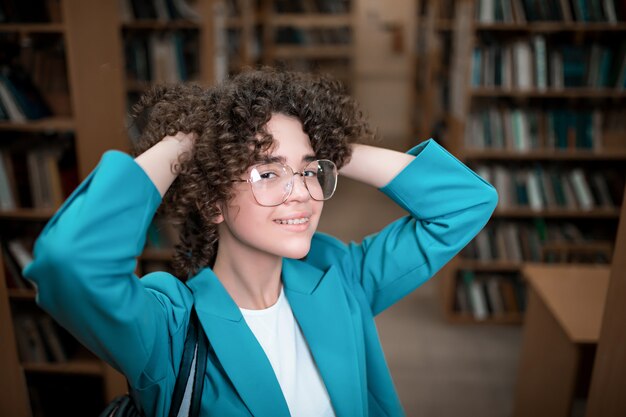 Giovane bella ragazza riccia in bicchieri in una biblioteca