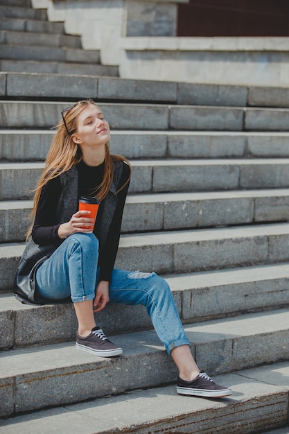 Giovane bella ragazza seduta sui gradini con caffè in bicchieri.