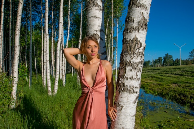 Giovane bella ragazza snella in un lungo vestito di corallo che posa in un boschetto di betulle che sorride con il suo petto nudo