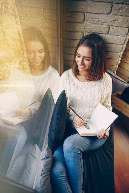 Giovane bella ragazza sorridente in maglione bianco e jeans che si siedono sul davanzale della finestra e che disegnano un'immagine nello sketchbook