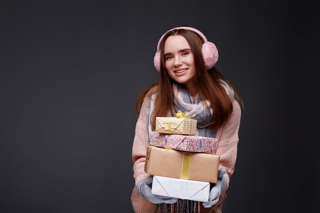 Giovane bella ragazza sorridente in maglione lavorato a maglia e paraorecchie lanuginose rosa che tengono i contenitori di regalo