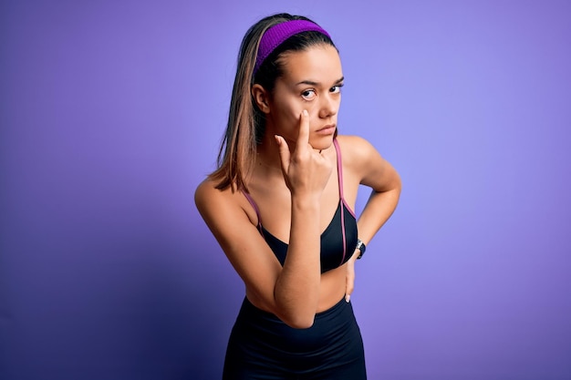 Giovane bella ragazza sportiva che fa sport indossando abbigliamento sportivo su sfondo viola isolato Indicando l'occhio che ti guarda gesto espressione sospetta