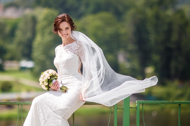 Giovane bella ragazza sposa con bouquet da sposa di rose si siede vicino al fiume il vento soffia il velo