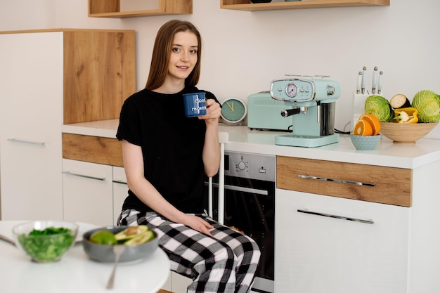 Giovane bella ragazza vegetariana vestita in pigiama che mangia frutta e verdura per colazione a casa in cucina