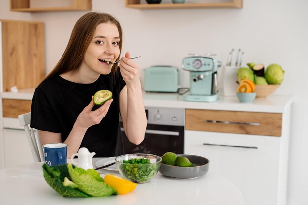 Giovane bella ragazza vegetariana vestita in pigiama che mangia frutta e verdura per colazione a casa in cucina