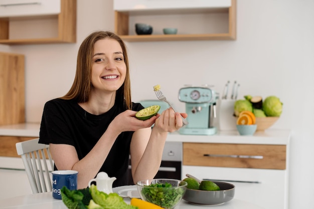 Giovane bella ragazza vegetariana vestita in pigiama che mangia frutta e verdura per colazione a casa in cucina