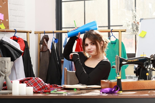 Giovane bella sarta seduta all'atelier e tenendo il suo taccuino mentre guarda la telecamera