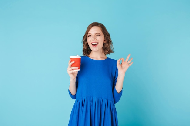 Giovane bella signora in abito in piedi con una tazza di caffè su sfondo blu. Bella ragazza che mostra felicemente il gesto giusto mentre guarda nella fotocamera