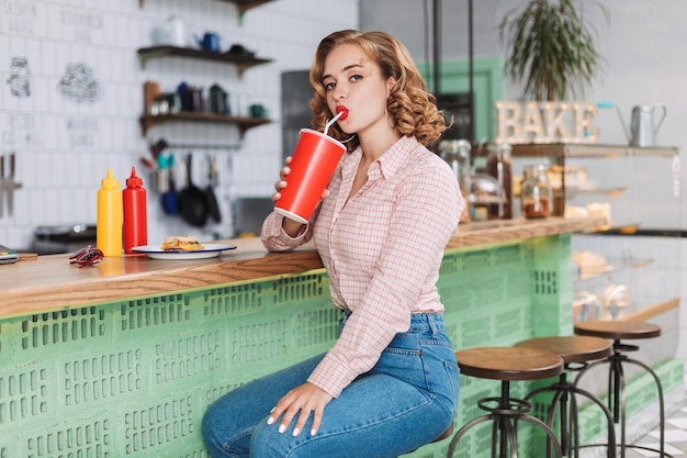 Giovane bella signora in camicia e jeans seduta al bancone del bar e bere acqua gassata mentre guarda cupamente a porte chiuse nella caffetteria