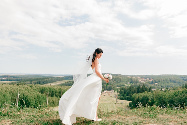 Giovane bella sposa in vestito da sposa bianco che posa su una collina alla natura.