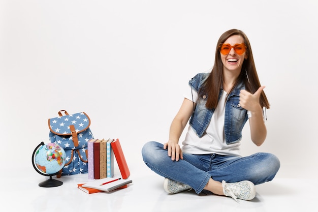 Giovane bella studentessa allegra con gli occhiali a cuore rosso che mostra il pollice seduto vicino ai libri di scuola dello zaino del globo isolati