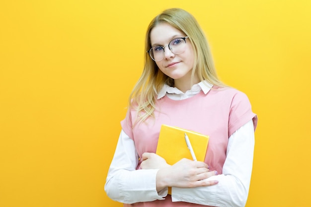 Giovane bella studentessa bionda con gli occhiali tiene libri e una penna su uno sfondo giallo isolato