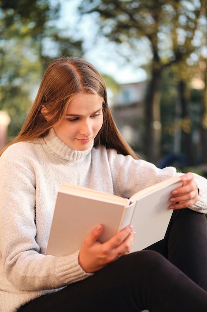 Giovane bella studentessa in maglione accogliente che legge sognante il libro mentre studia all'aperto