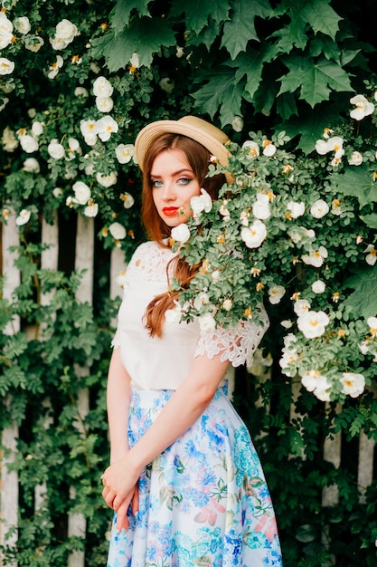 Giovane bellezza russa. Ragazza attraente in gonna lunga retrò vintage, top vecchio stile bianco e capelli rossi ricci e cappello di paglia in posa per la fotocamera con recinzione e alberi verdi