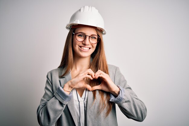 Giovane bellissima architetta dai capelli rossi donna che indossa un casco di sicurezza su sfondo bianco sorridendo innamorata mostrando il simbolo del cuore e la forma con le mani concetto romantico