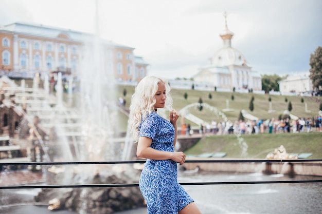 Giovane bellissima donna bionda con i capelli in abito blu che cammina vicino alle fontane