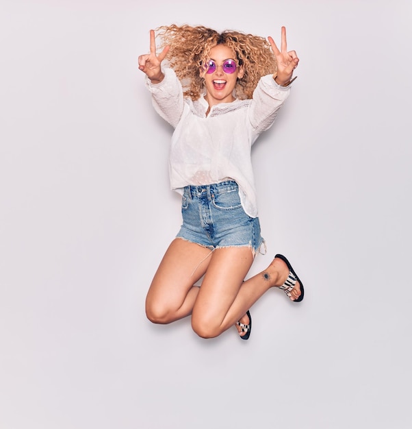 Giovane bellissima ragazza riccia che indossa abiti casuali e occhiali da sole retro sorridente felice saltando con il sorriso sul viso facendo il segno della vittoria su uno sfondo bianco isolato