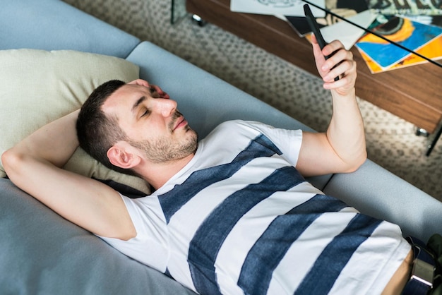 Giovane bello che guarda il suo smartphone mentre è sdraiato sul divano a casa