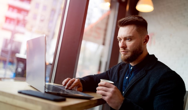 Giovane bello che lavora con il computer portatile in un caffè moderno Uomo alla moda che lavora con il notebook e beve caffè