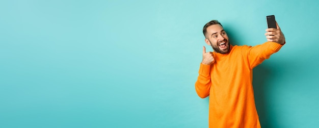 Giovane bello che prende selfie sul telefono cellulare che mostra i pollici in su alla registrazione della fotocamera dello smartphone vl