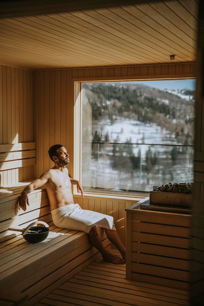 Giovane bello che si rilassa nella sauna e guarda la foresta d'inverno attraverso la finestra