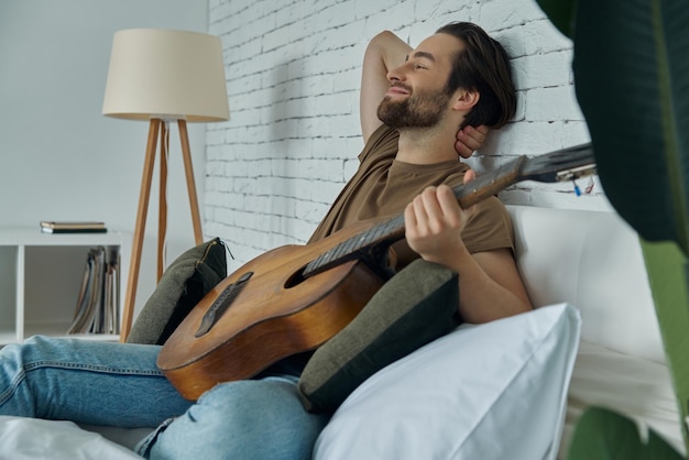 Giovane bello che tiene la chitarra acustica mentre si rilassa sul letto a casa