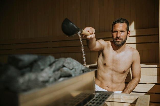 Giovane bello che versa acqua sulla pietra calda nella sauna