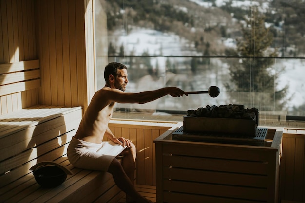 Giovane bello che versa acqua sulla pietra calda nella sauna