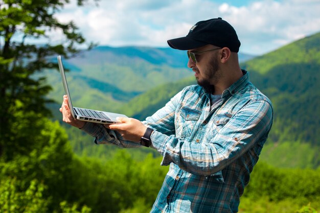 Giovane bello uomo alla moda che lavora al computer portatile nelle montagne all'aperto