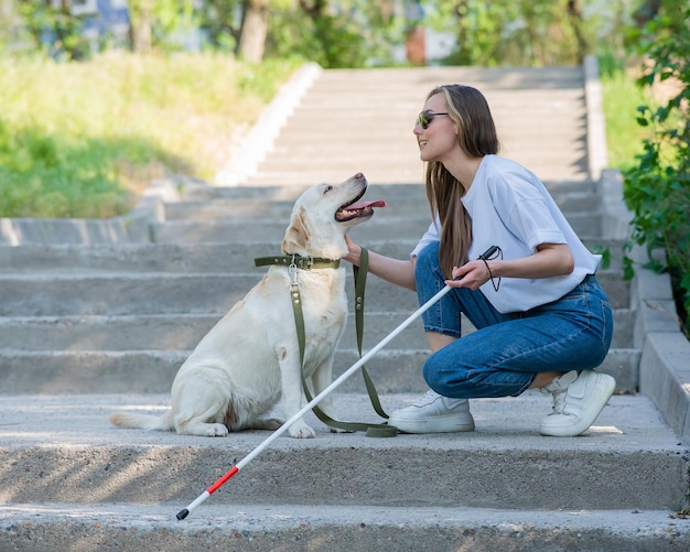 Giovane bionda cieca che accarezza un cane guida in una passeggiata nel parco donna con bastone tattile alle scale