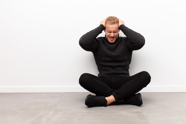 Giovane bionda sentendosi stressata e frustrata, alzando le mani alla testa, sentendosi stanca, infelice e con emicrania seduta sul pavimento di cemento