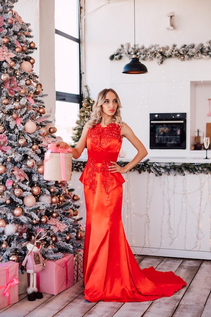 Giovane bionda sexy in un lungo vestito rosso vicino all'albero di Natale splendidamente decorato