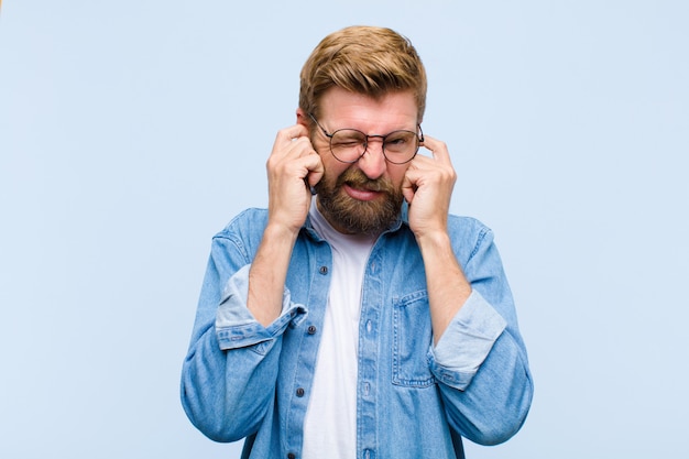 Giovane biondo adulto che sembra arrabbiato, stressato e infastidito, coprendo entrambe le orecchie con un rumore assordante, suono o musica ad alto volume