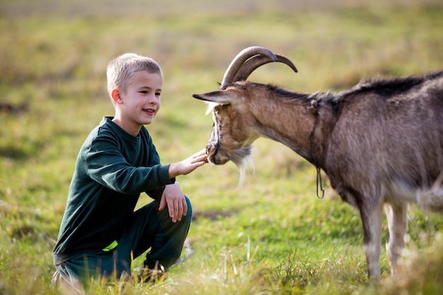 Giovane biondo carino bel ragazzo sorridente bambino che gioca con la capra cornuta barbuta all'aperto in estate soleggiata luminosa o primavera su sfocato verde chiaro erboso