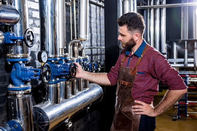 Giovane birraio maschio in grembiule in pelle che supervisiona il processo di fermentazione della birra nella moderna fabbrica di birra fa...