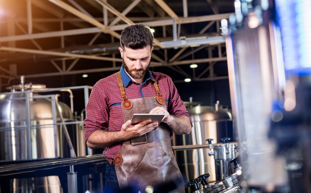 Giovane birraio maschio in grembiule in pelle che supervisiona il processo di fermentazione della birra nella moderna fabbrica di birra fa...